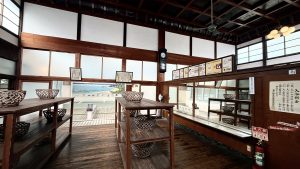 This photo was taken at the Edo-Tokyo Open Air Architectural Museum in Koganei, Tokyo. It shows the interior of a traditional Japanese public bathhouse (sento) with furniture like a table, baskets, a ceiling fan, a ceiling light, fire extinguisher.