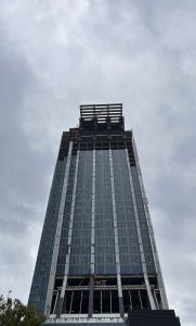A tall skyscraper under construction, viewed from below, with a cloudy sky in the background.