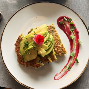 A savory corn fritter is topped with sliced avocado, a small pink edible flower, and fresh herbs, served on a white plate with a decorative swipe of bright pink sauce and microgreens.