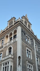 Corner view of a historic building in Prague with detailed sgraffito artwork, arched balconies, and stone statues. The facade combines beige brick and ornate white decorations under a clear blue sky.