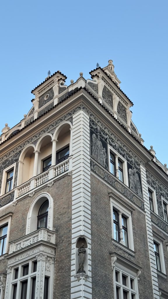 Corner view of a historic building in Prague with detailed sgraffito artwork, arched balconies, and stone statues. The facade combines beige brick and ornate white decorations under a clear blue sky.