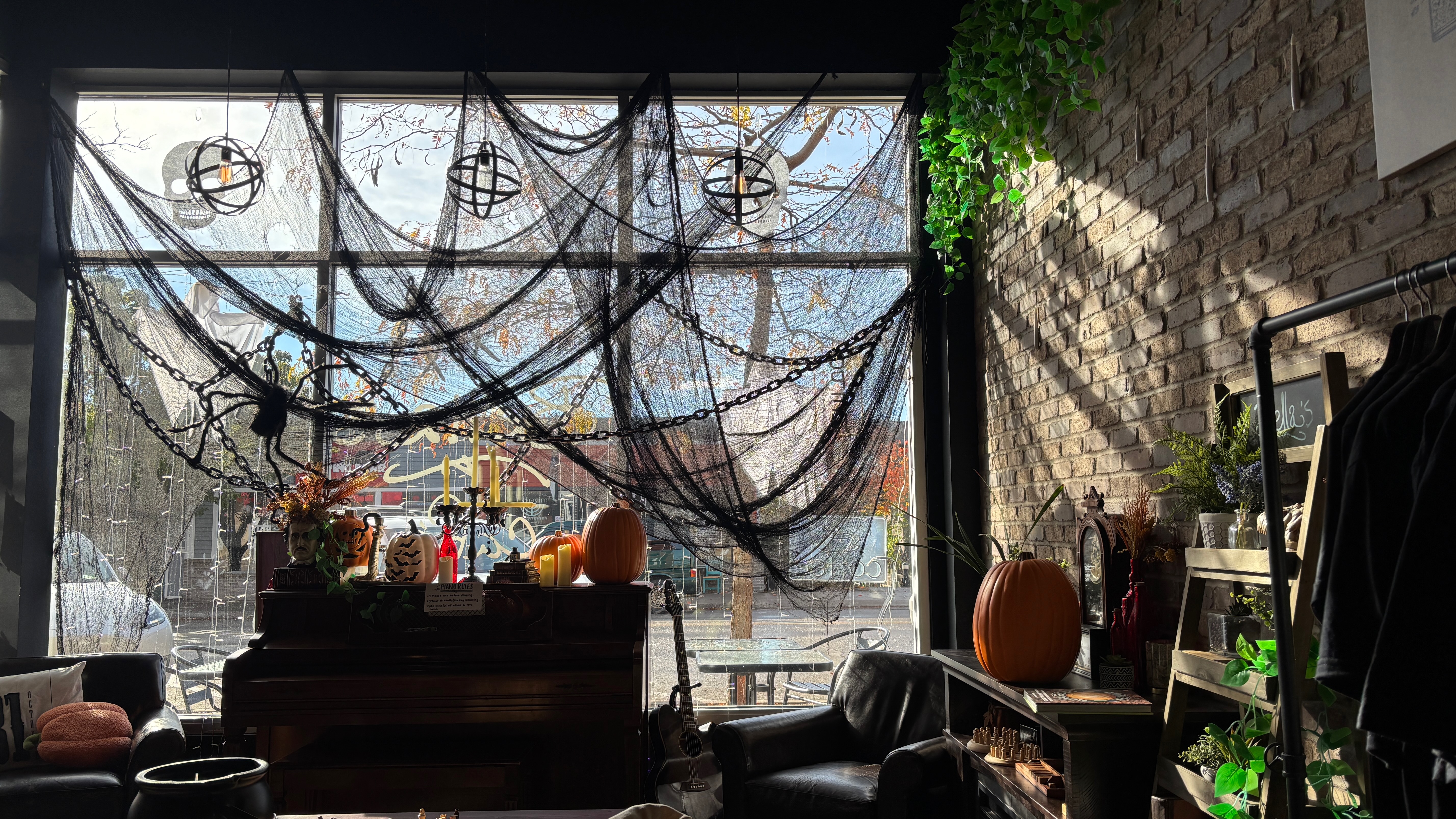 From the inside, a storefront decorated for halloween. There's a large window with a piano in front. There are fake spider webs hanging in the window and there are many pumpkins.