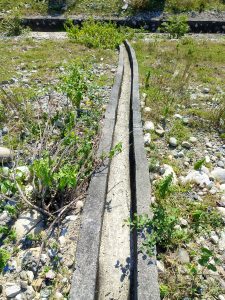 A narrow, concrete ditch runs through a grassy landscape, flanked by small shrubs and patches of greenery.