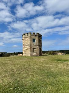 A small, circular stone tower with battlements stands on a wide grassy field under a bright blue sky with scattered clouds.