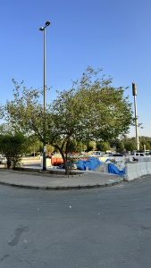 A roundabout scene in an outdoor area featuring a small tree on the left side, alongside a tall streetlight. In the background, piles of materials covered with blue tarps are visible, as well as some parked vehicles.