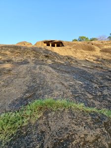 A view of the Kanheri Caves carved into a rocky hill in Borivali, Mumbai. The ancient cave entrance sits above the wide stone slope under a bright blue sky.