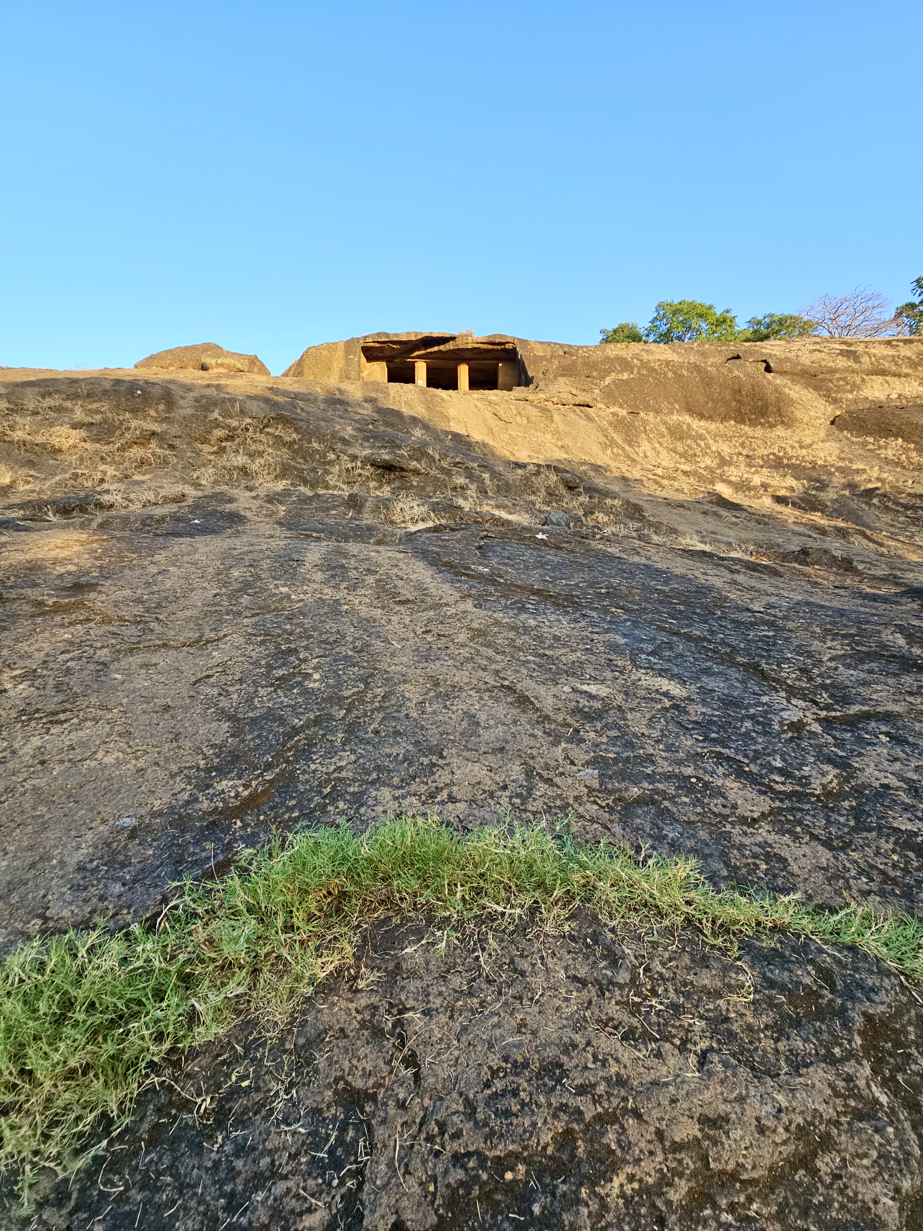A view of the Kanheri Caves carved into a rocky hill in Borivali, Mumbai. The ancient cave entrance sits above the wide stone slope under a bright blue sky.