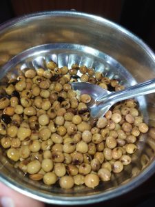 A close-up image of a stainless steel bowl filled with light brown and tan-colored dried legumes, some of which show darker spots. 
