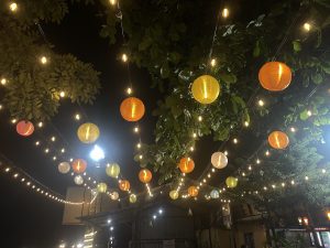 A nighttime scene featuring colorful paper lanterns and warm string lights hanging from above, surrounded by lush green leaves