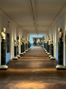 The Corridoio Vasariano in Florence, lit with warm golden light, with marble busts on green columns along the walls.
