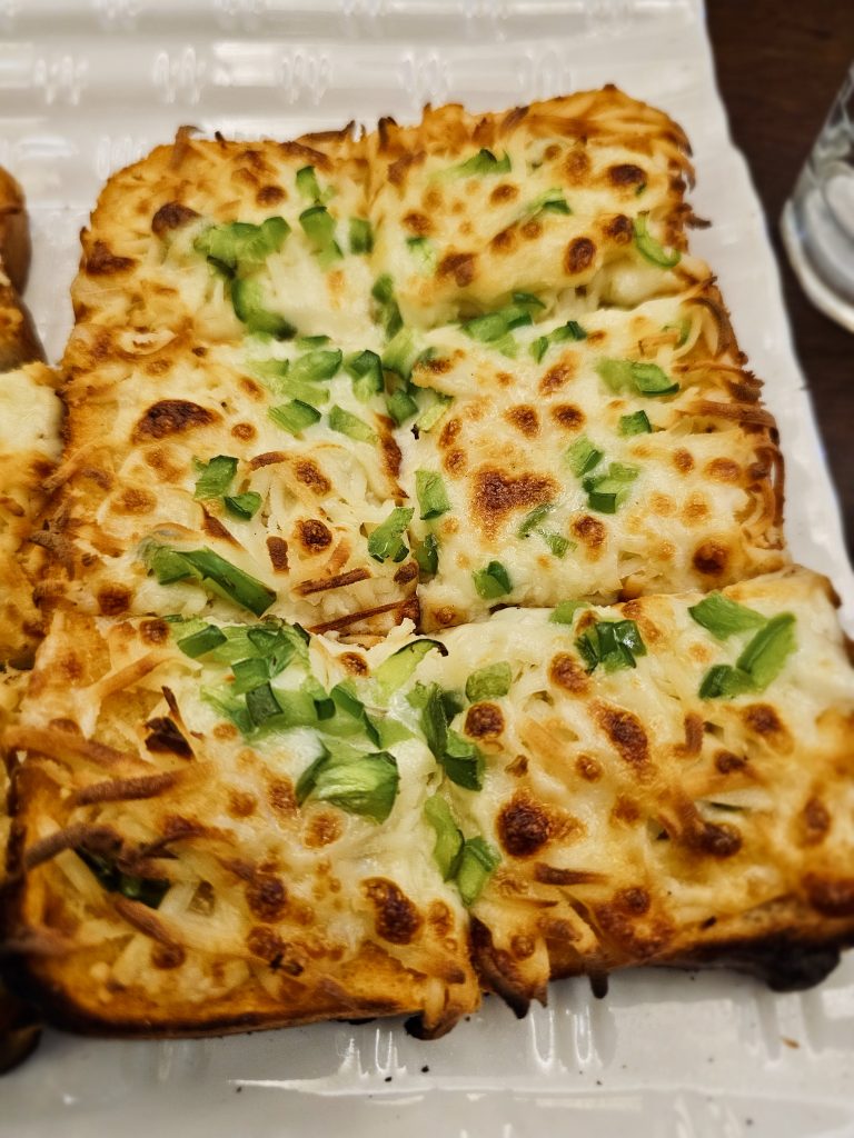 A plate of golden, cheesy garlic bread topped with green capsicum, captured near a cafe in Malabar Hill, Mumbai. The melted cheese and crisp edges make it look warm and tasty.