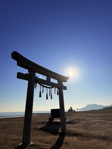 A seaside torii gate under a clear blue sky, with the rising sun over a calm ocean and a distant cape.