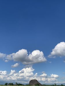 A distant rock formation resembling a small hill beneath a bright blue sky with large white clouds, surrounded by flat green fields.
