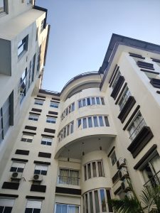 A view of a modern multi-story building from a low angle, showcasing a curved section at the top, large windows, and a mix of white and brown exterior elements