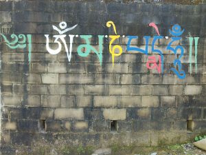 Colorful Tibetan script painted on a weathered brick wall. The text features the mantra “Om Mani Padme Hum” written in stylized calligraphy, with each syllable in a different color—white, green, yellow, blue, red, and turquoise. The wall shows signs of age and moisture, with dark stains and patches of moss near the base.