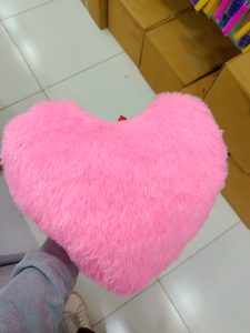 A person holding a large, fluffy pink heart-shaped pillow in a store, with cardboard boxes visible in the background and a tiled floor.