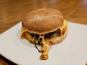 A tasty burger on a white plate, filled with melted cheese, vegetables, chicken, and a soft bun. The cheese is spilling out, making it look delicious and warm. Captured from a coffee stored in Kozhikode, Kerala.  