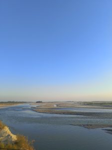 A serene landscape featuring a wide river meandering through a sandy, rocky delta under a clear blue sky.
