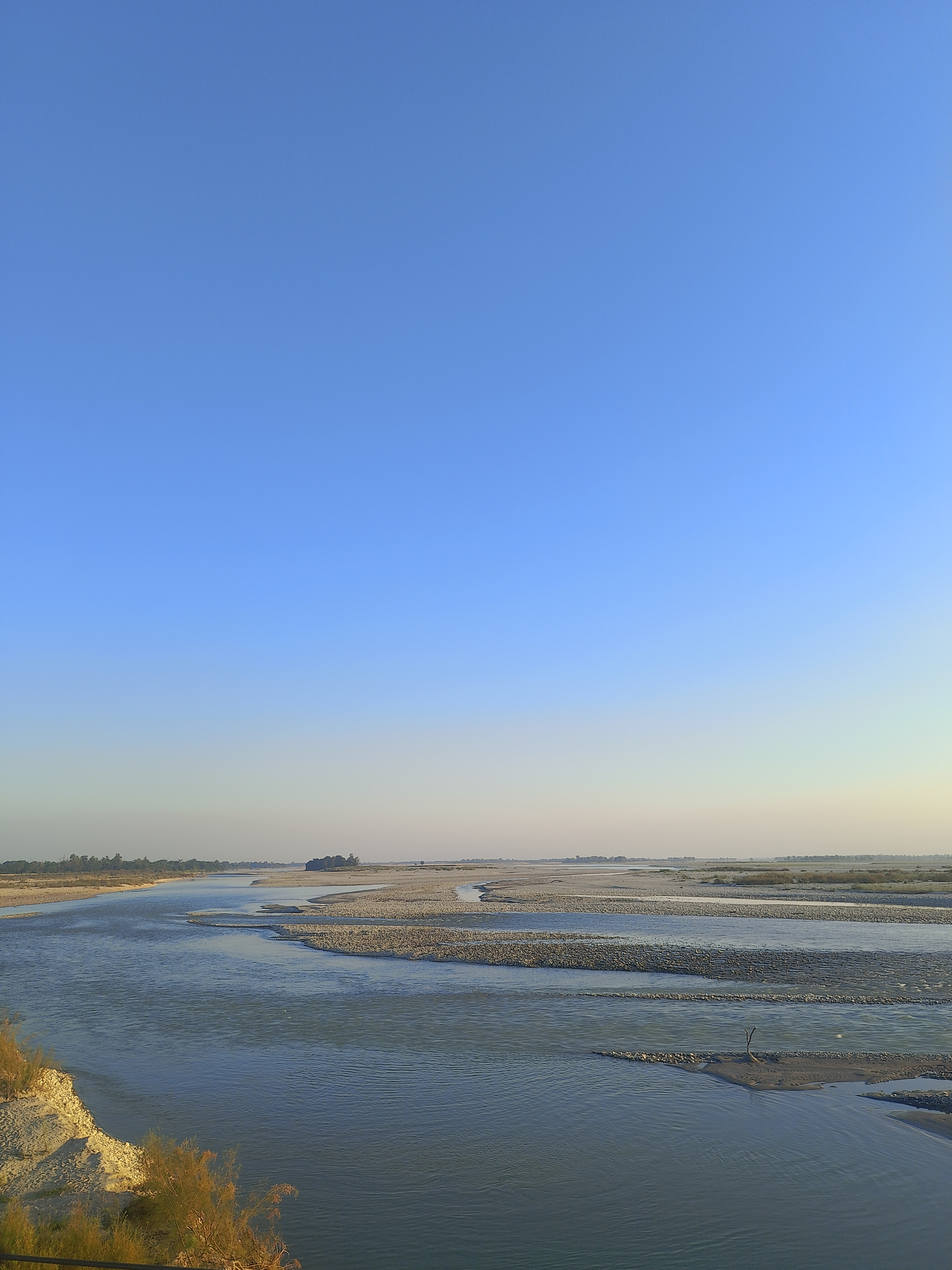 A serene landscape featuring a wide river meandering through a sandy, rocky delta under a clear blue sky.