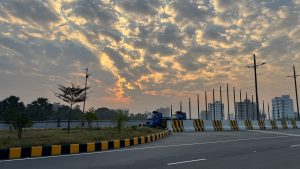 Golden rays of sunlight burst through the clouds above a wide city street with parked blue trucks.
