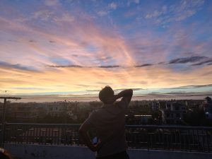 A person stands on a rooftop, gazing at a vibrant sunset. 