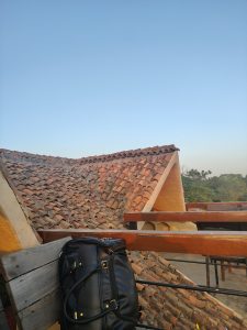 

A rooftop with terracotta tiles under a clear blue sky, a black bag on the railing, and greenery in the background.