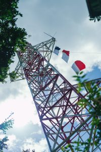 A dramatic low-angle shot looking up at a towering red and white steel lattice structure, possibly a transmission tower or radio mast. The geometric metal framework is framed naturally by green foliage in the foreground. At the top, wind indicators flutter against a bright blue sky scattered with soft white clouds.