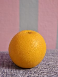A bright orange is placed on a textured cloth surface with a pastel pink and blue striped wall in the background, captured indoors at Perumanna, Kozhikode, Kerala, showcasing the natural texture and color of the fruit.