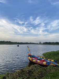 A vibrant, flower-adorned boat is docked by a serene lake under a bright blue sky with wispy clouds. Another boat floats in the distance. Tranquil and picturesque.