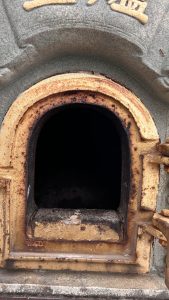 Close-up of a small furnace-like opening with a rusty yellow frame and dark charred interior.
