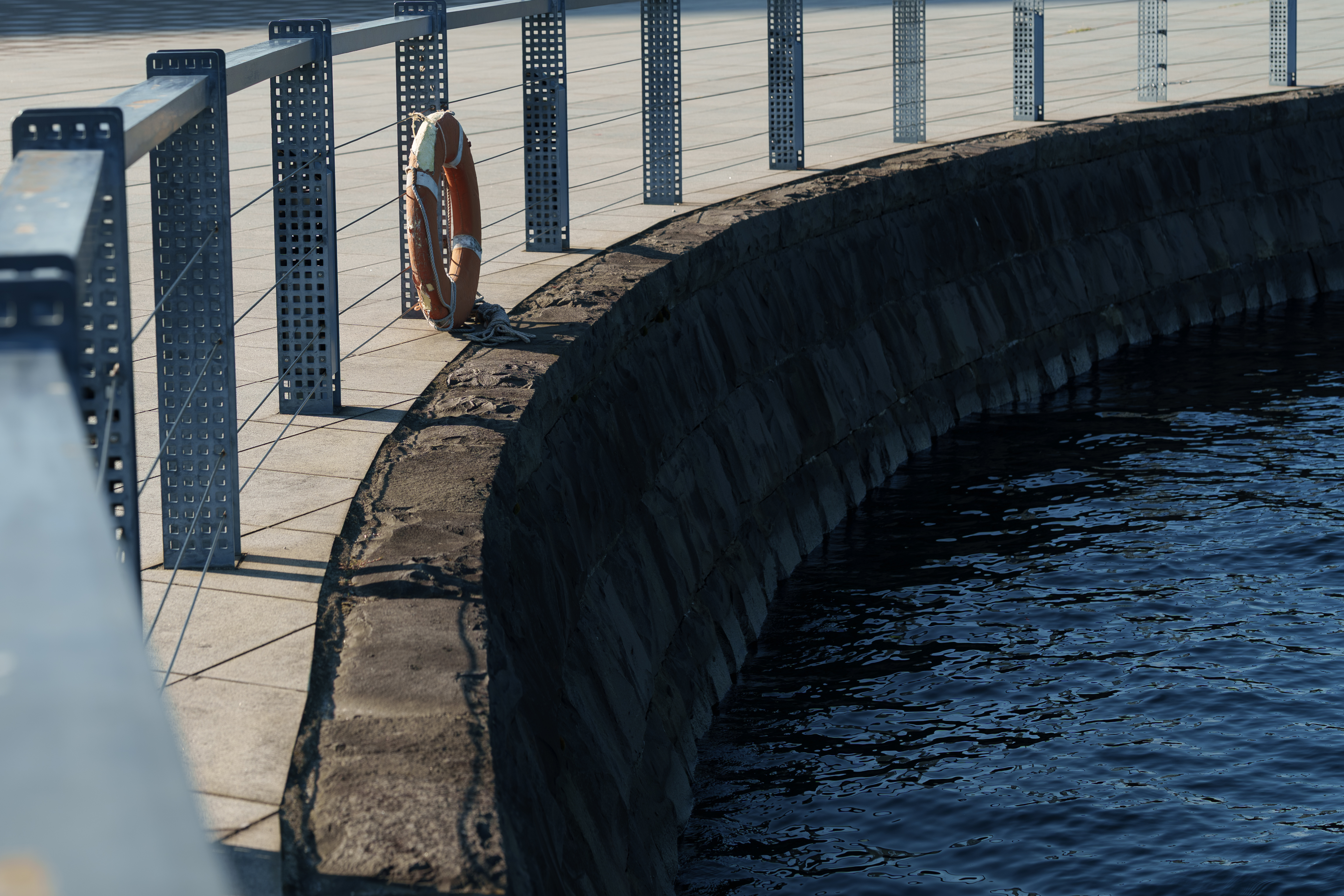 A lifebuoy hangs along a curved waterfront with a stone wall and steel railing. The calm, dark blue water reflects the soft afternoon light.