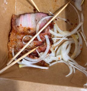 A partially eaten meal in a brown cardboard container, featuring a piece of cooked pork with visible layers of fat and seasoning.