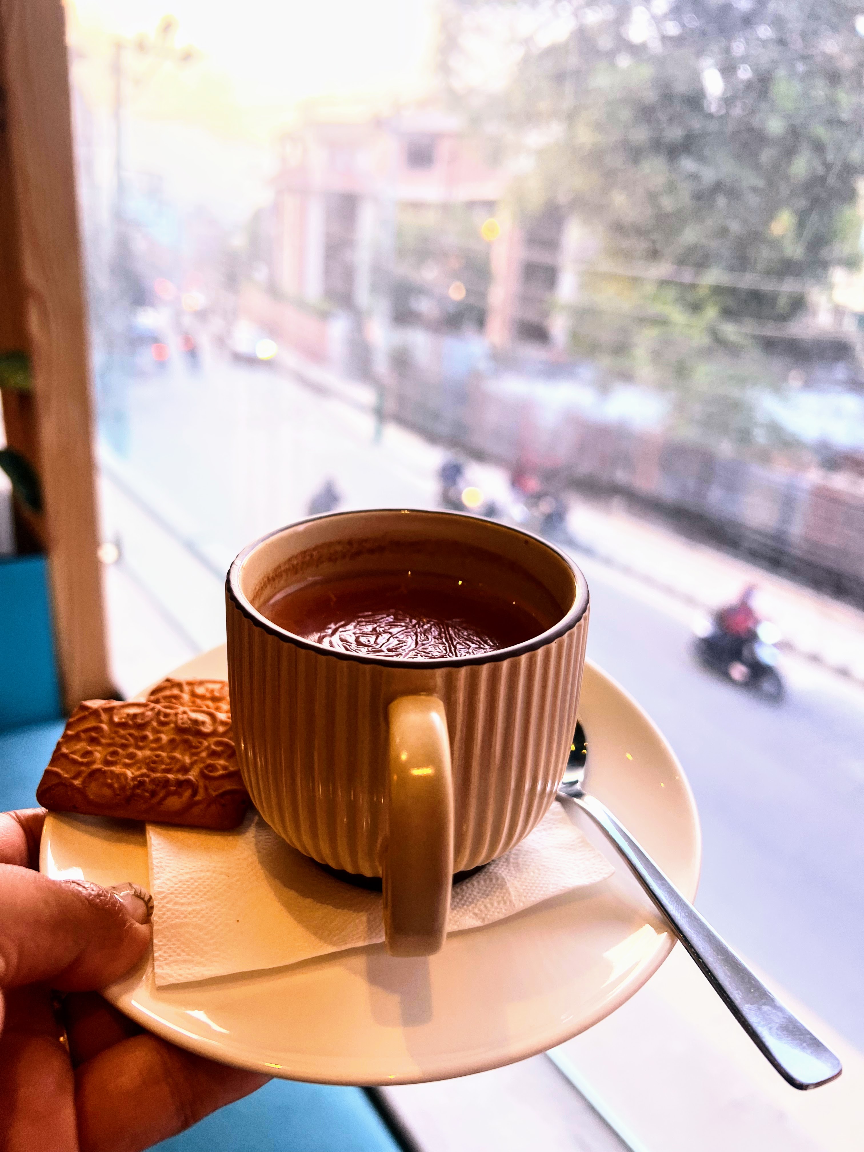 A hand holds a beige ribbed mug of hot dark tea on a saucer with a spoon and two biscuits, viewed from an indoor window looking out onto a blurred street with traffic.
