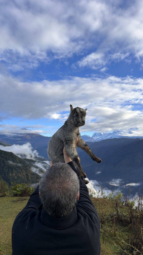 A person holding a small goat in the air against a backdrop of mountains and a cloudy blue sky.