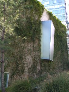 A modern, partially covered building facade is adorned with lush green vegetation, featuring a large, frosted glass box protruding from the wall. 