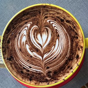 A close-up, overhead view of a hot chocolate or mocha in a yellow-rimmed cup featuring detailed latte art of a white heart shape with concentric rings, dusted with cocoa.