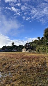 A scenic landscape featuring a large open field with patches of dry grass, surrounded by hills and buildings in the background. 