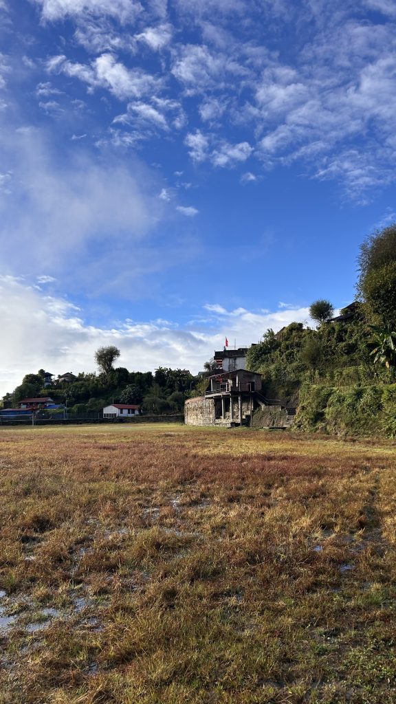 A scenic landscape featuring a large open field with patches of dry grass, surrounded by hills and buildings in the background.