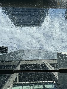 A picture from a rainy day, from under the glass roof top with raindrops on the glass. The behind-glass view has some tall buildings and a cloudy sky.