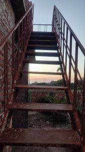 A low-angle view looking up a rusty red iron staircase attached to a rough brick wall. Through the open steps, a soft sunset sky with orange hues and distant trees is visible.