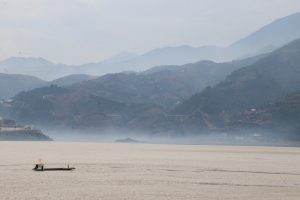 A misty, mountainous landscape over a wide body of water. In the foreground, the water is calm, and a small, simple boat carrying at least one person is crossing the lower part of the frame. In the distance, a thick layer of fog or mist hugs the waterline, partially obscuring the base of the hills. The picture focuses on a boat with the number 03, and a man wearing an orange coloured jacket.
