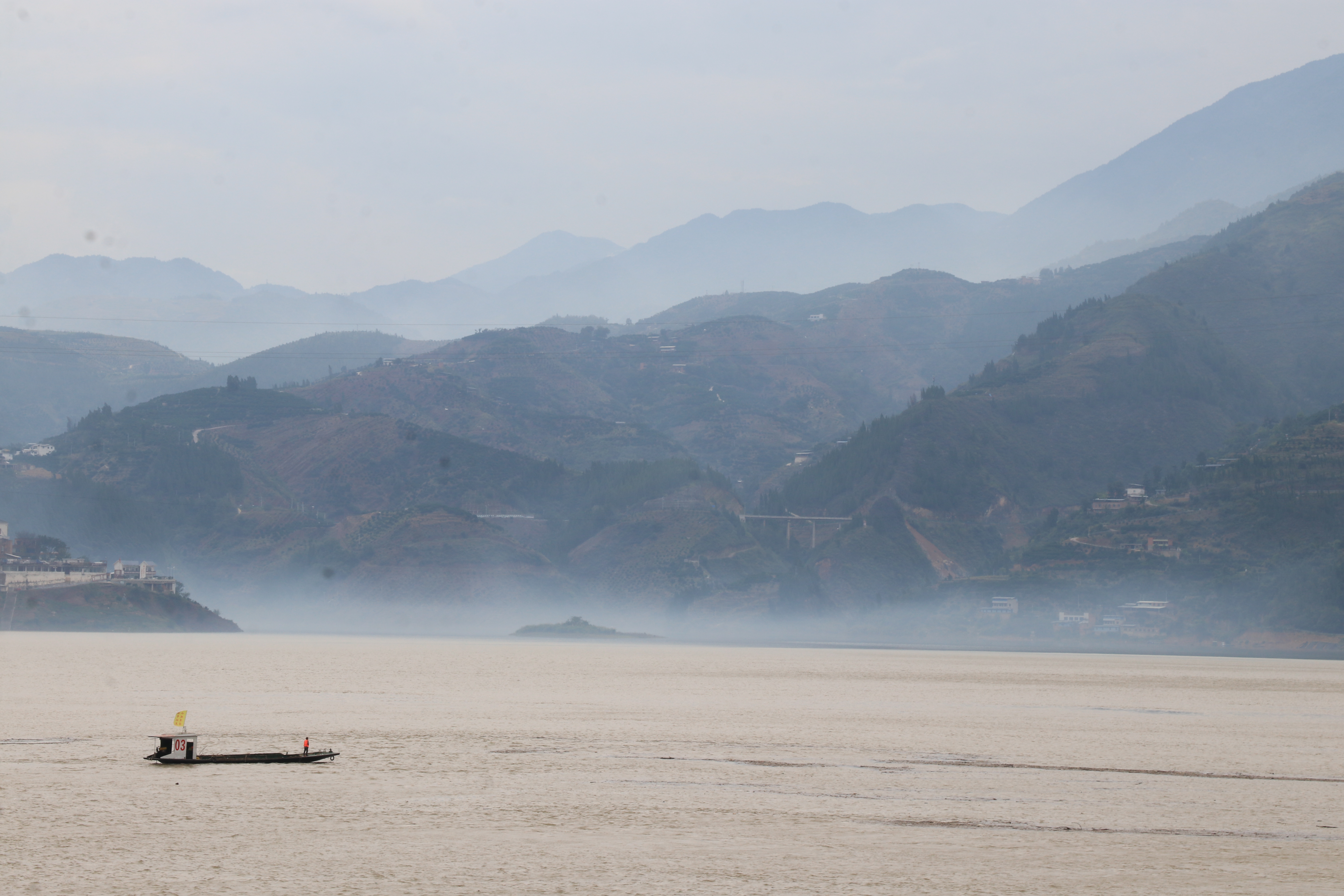 A misty, mountainous landscape over a wide body of water. In the foreground, the water is calm, and a small, simple boat carrying at least one person is crossing the lower part of the frame. In the distance, a thick layer of fog or mist hugs the waterline, partially obscuring the base of the hills. The picture focuses on a boat with the number 03, and a man wearing an orange coloured jacket.