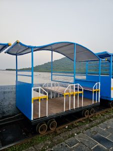 A small blue open coach of a mini train on narrow tracks near the water. Hills and mangroves are visible in the background. Captured near Elephanta Caves, Mumbai, Maharashtra.  