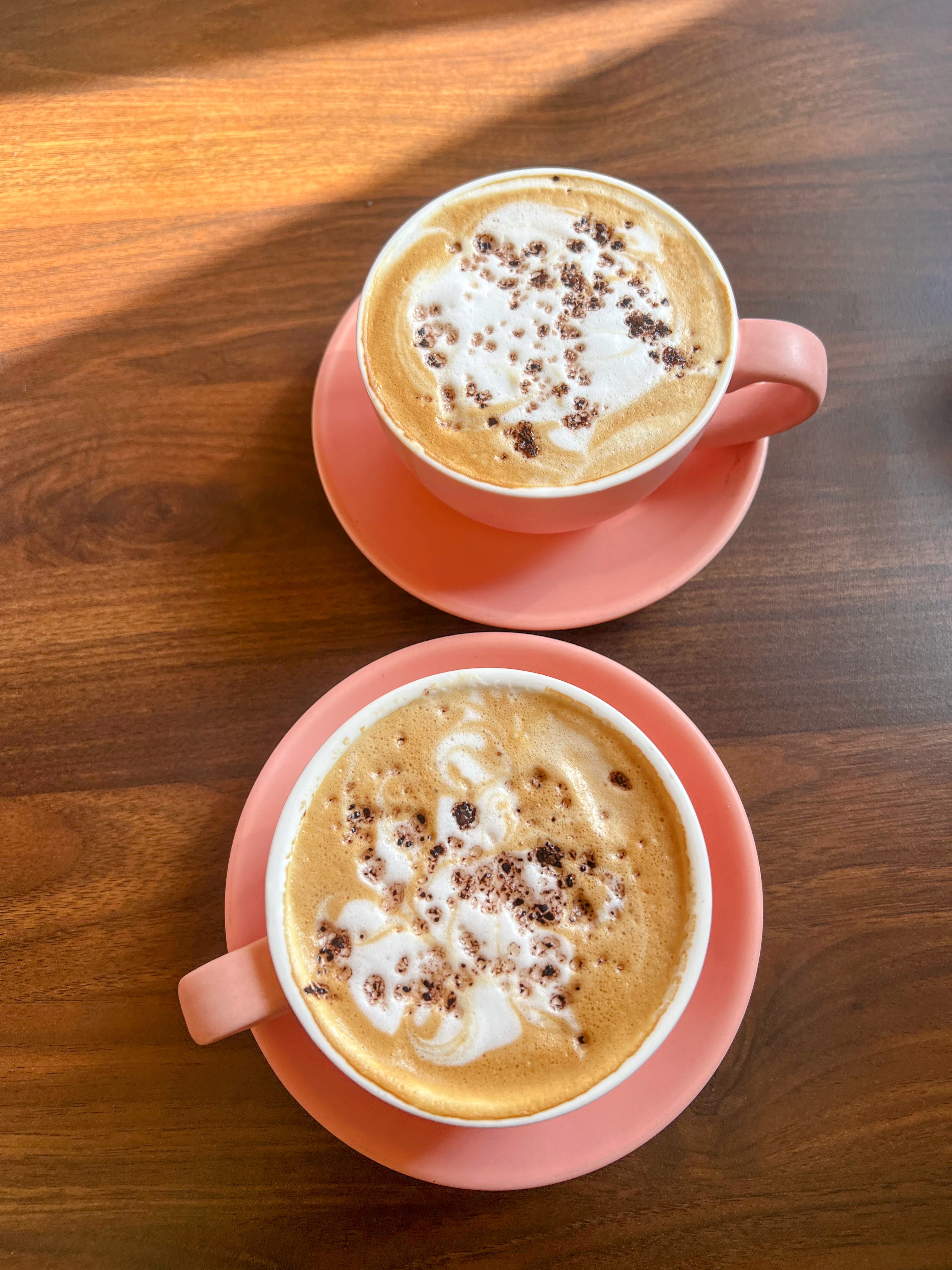 Two identical cappuccinos, served in small pink cups on matching saucers, sit on a dark wooden table with natural light hitting the top cup.