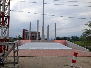 A tall gray-column monument on a brick platform with a red-brick backdrop, a nearby building, and a field under a cloudy sky in Tetulia, Panchagarh.