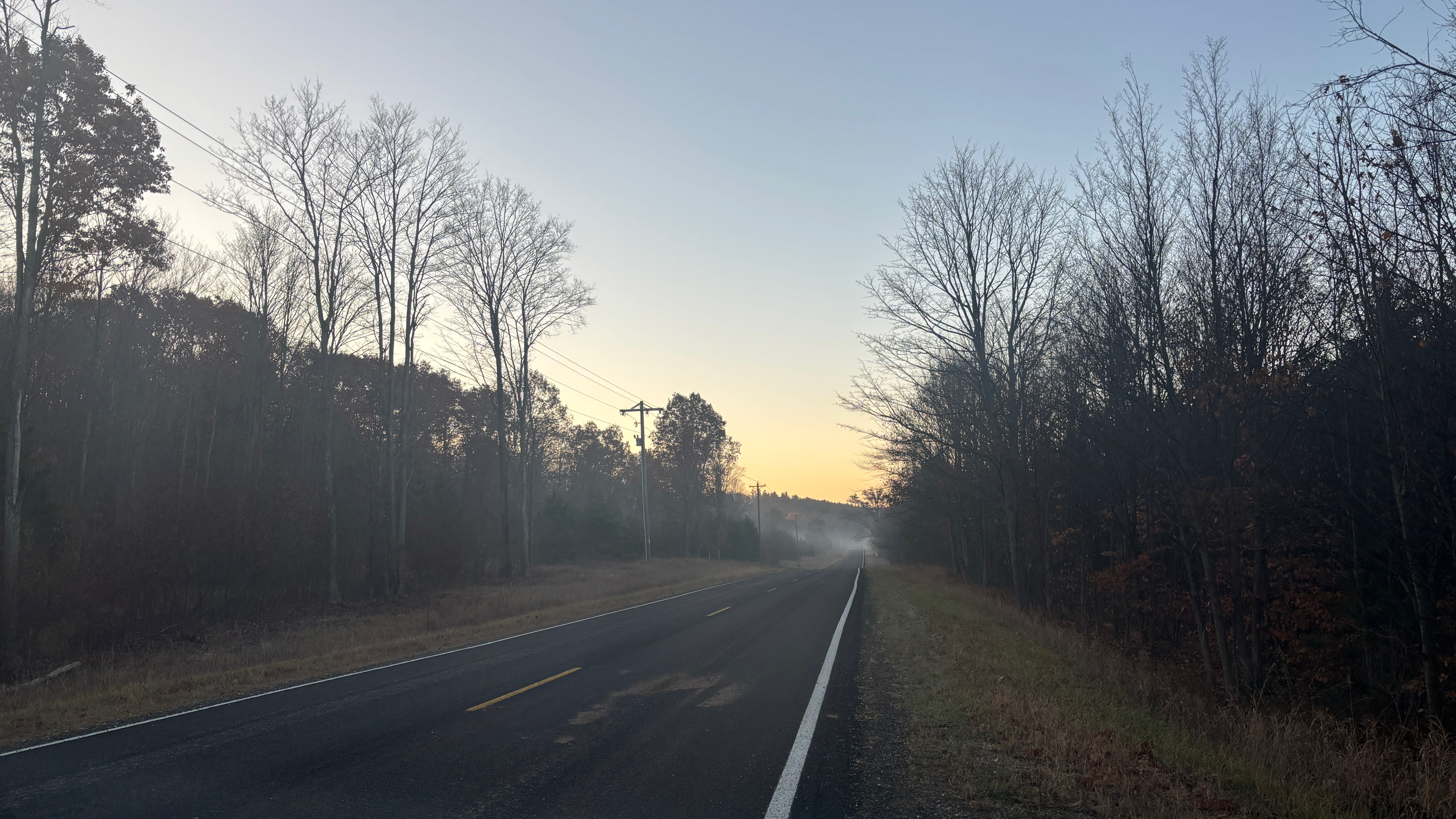 A long straight road made of blacktop goes off into the distance. It's lined by forest. In the distance the road is obscured by mist. The sky is lit by morning light.
