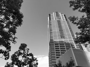 A black and white skyscraper from a low angle. The building features sleek glass windows and a mix of architectural designs. Surrounding the base are various leafy trees in a monochrome scene.