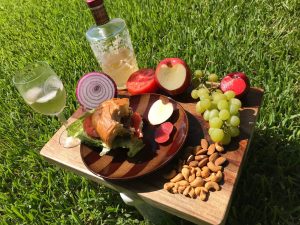 A breakfast beautifully decorated and placed on a brown table and a brown plate. The plate contains half a burger, and the table contains half a sliced onion, apple, tomato, peach, grapes, almond nuts, and a glass of wine with its bottle.