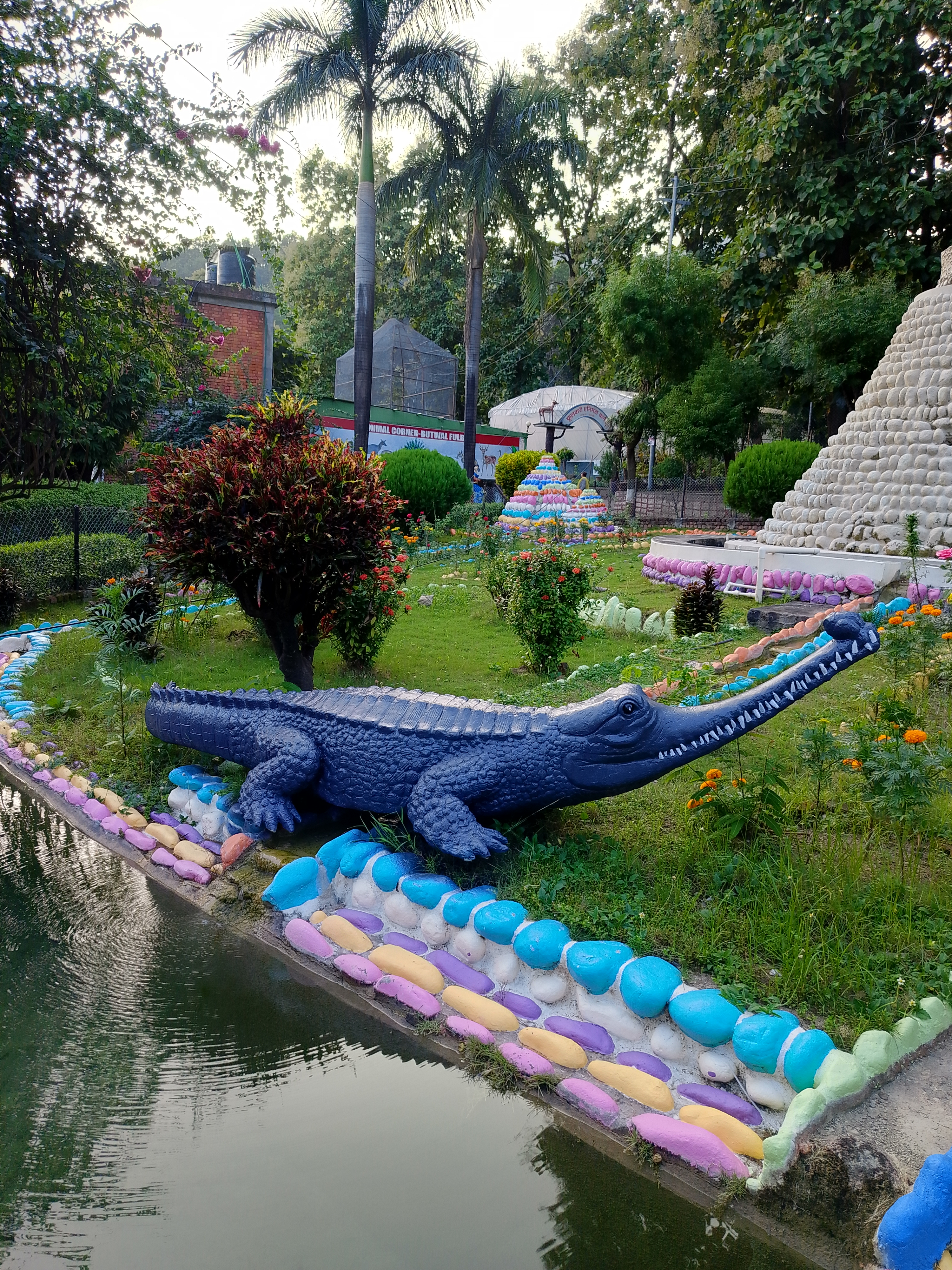 A large, colorful crocodile sculpture is positioned on a grassy bank beside a small pond.