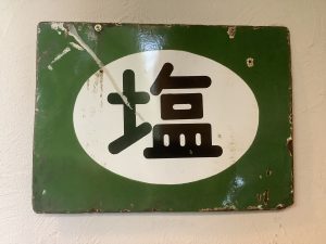 Vintage Japanese salt signboard made of enamel painted metal. The metal is battered and rusty in places, sign is fixed with two top screws to a rendered wall.